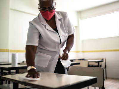 A masked woman cleans a school desk with a spray bottle and cloth