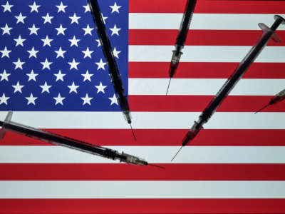 Medical syringes seen in front of the national flag of the United States of America, displayed on a screen in the background.
