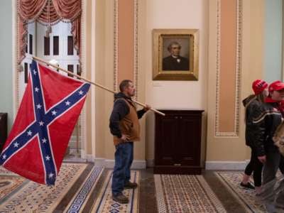 A white man, inexplicably still alive after storming the U.S. capitol building, holds the flag of a defunct seditionist government