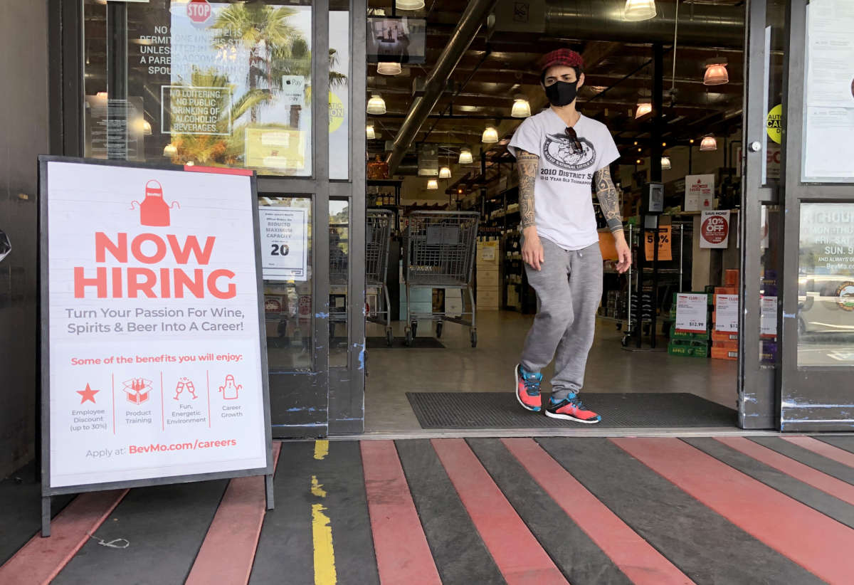 A man exits a store with a "NOW HIRING" sign in front of it