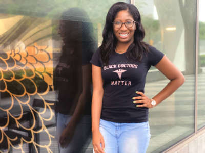 A Black woman wearing a shirt reading "BLACK DOCTORS MATTER" stands, smiling, with one hand on her hip.