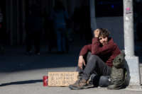 An unhoused person sits on the sidewalk with a cardboard sign reading "Have a great day and stay positive" during a pandemic of the novel coronavirus