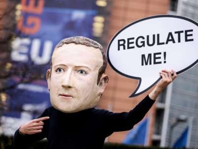 A protester in a deeply upsetting paper mache mask of Mark Zuckerberg holds a sign resembling a word bubble, with the text "REGULATE ME" written within