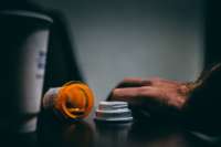 a photo of a pill bottle spilling out onto a surface with someone's hand in the right side of the frame and a coffee cup on the left