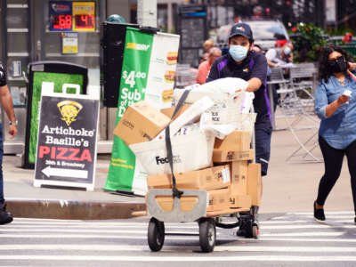 A masked fed-ex worker pushes a dolly stacked with packages