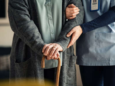 Elder woman using walking cane at nursing home with nurse holding hand for support.
