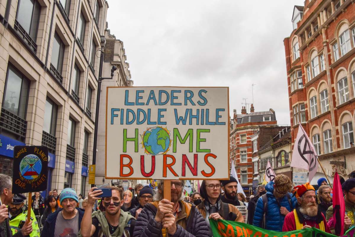 Unmasked people march in protest as one holds a sign reading "LEADERS FIDDLE WHILE HOME BURNS"