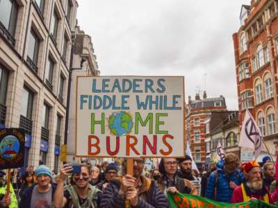 Unmasked people march in protest as one holds a sign reading "LEADERS FIDDLE WHILE HOME BURNS"