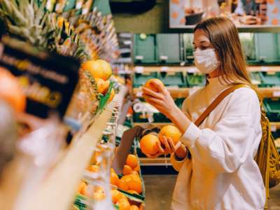 a masked woman holds several oranges in a grocery store.