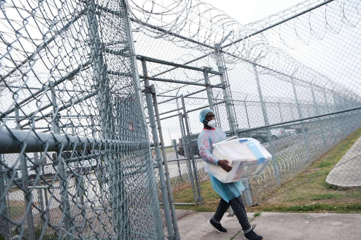 A medical worker walks a cooler of supplies through barbed wire gates and into a prison