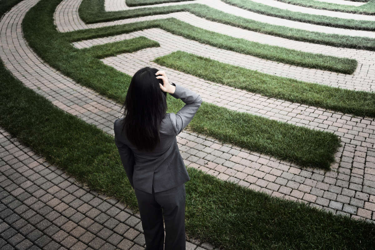 Young woman looking at lawn maze with hand on head, expressing uncertainty