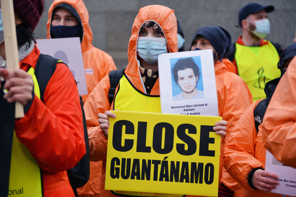 A protester holds a placard reading "Close Guantanamo" and portrait of detainee during an outdoor demonstration.