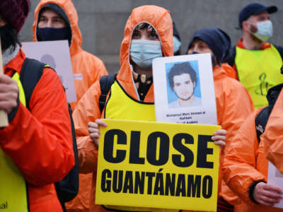 A protester holds a placard reading "Close Guantanamo" and portrait of detainee during an outdoor demonstration.
