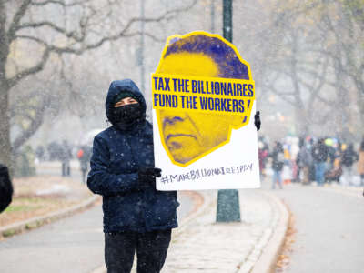 A masked protester holds a sign reading "TAX THE BILLIONAIRES; FUND THE WORKERS"