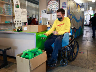 A person in a wheelchair tosses a green bag containing an overdose kit into a box filled with other, identical green bags