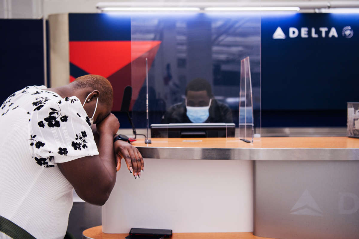 A woman rests her head in exhaustion on the counter of a Delta Airlines kiosk, where a masked employee works on a computer