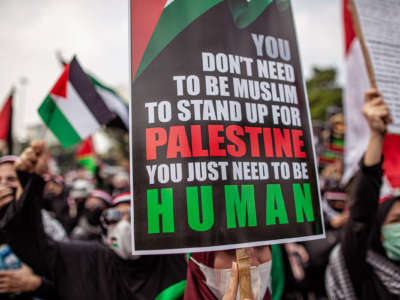 A protester holds a sign reading "YOU DON'T HAVE TO BE MUSLIM TO STAND UP FOR PALESTINE; YOU JUST NEED TO BE HUMAN"