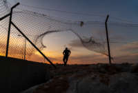 A person in silhouette runs through a torn fence