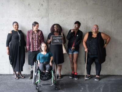 Six disabled people of color smile and pose in front of a concrete wall. Five people stand in the back, with the Black woman in the center holding up a chalkboard sign reading 'disabled and here.' A South Asian person in a wheelchair sits in front.