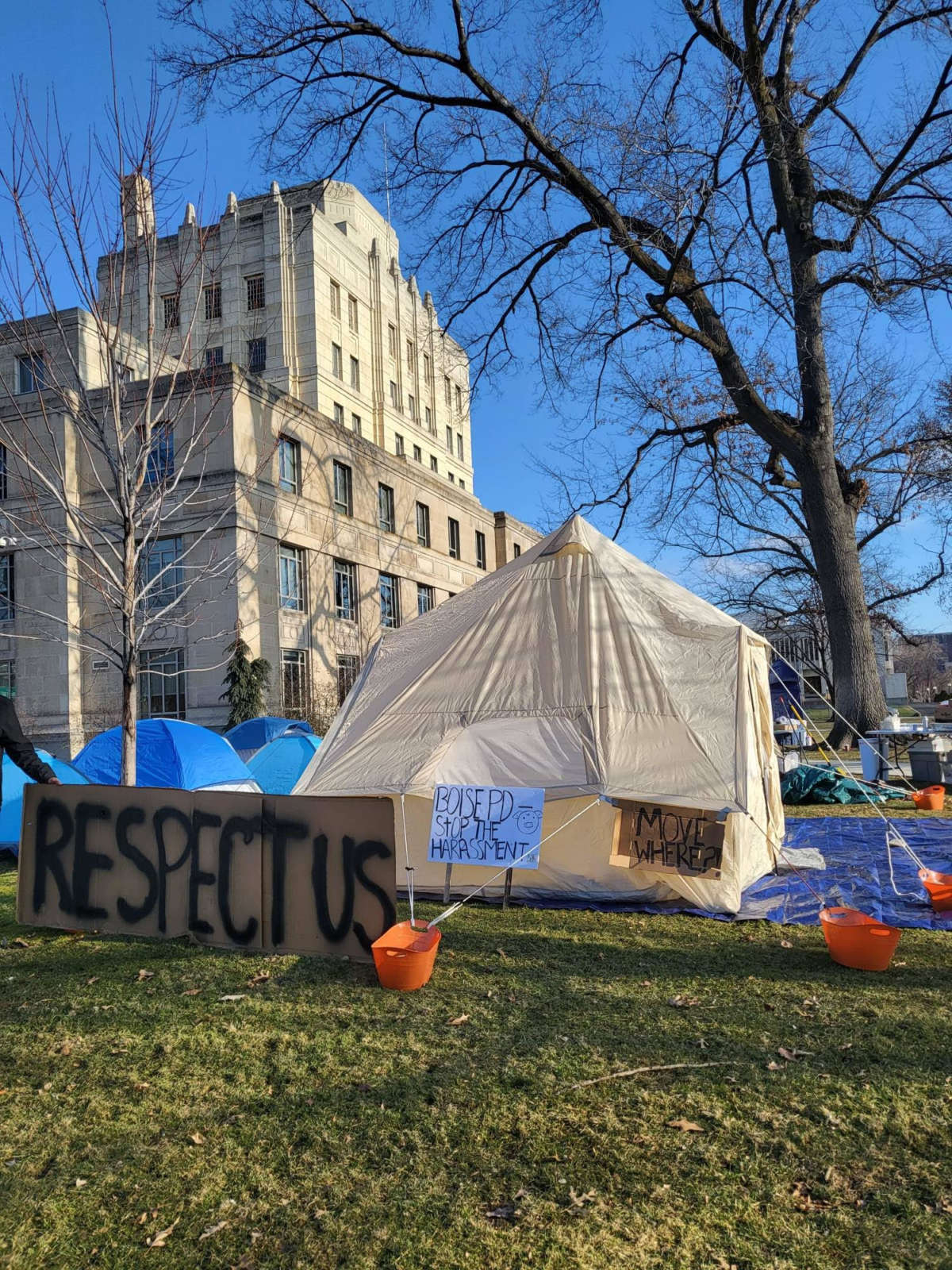 An Idaho government building is visible in the background of tents and signs demanding an end to harassment of the unhoused.