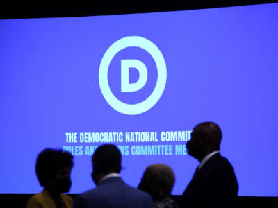 People walk in front of a projection screen showing the DNC logo