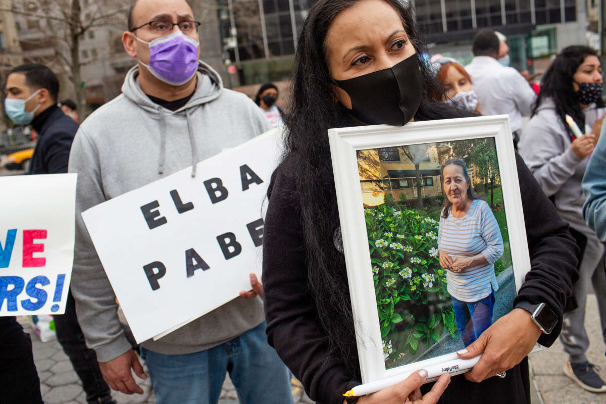 A masked woman at an outdoor protest holds a framed photo of an elderly loved one