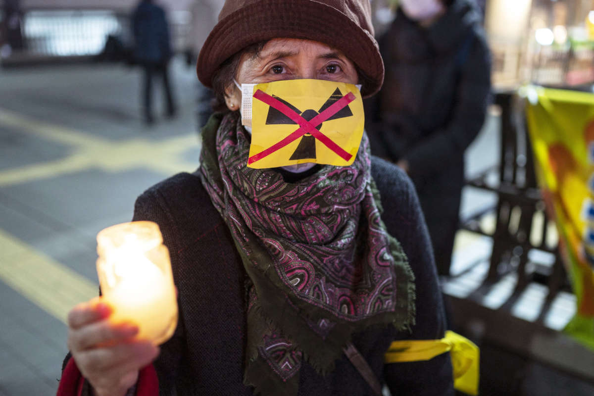 An elderly woman holds a candle while wearing a crossed-out nuclear power symbol over her medical face mask