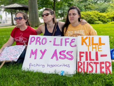 Three people embrace behind signs, one of which reads "PRO-LIFE MY ASS, FUCKING HYPOCRITES"