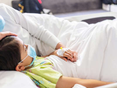 Mother with protective face mask holding son's hand while lying on the hospital bed with him.