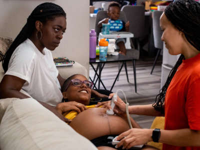Two black lesbians, one of whom is pregnant, hold eachother while a midwife performs an ultrasound exam