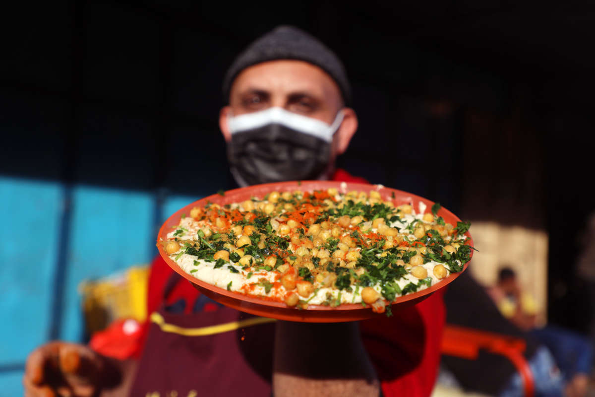 A masked man holds a plate of chickpea-topped hummus