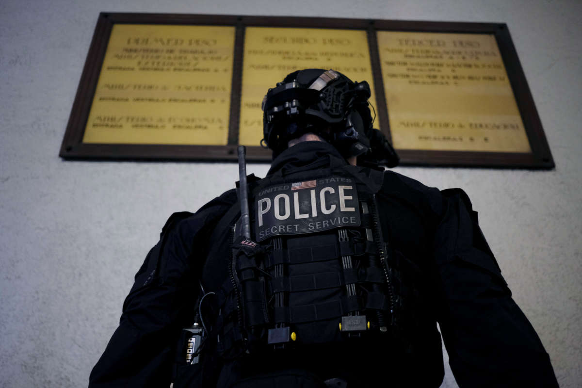 A secret Service member stands with his back towards the camera as he faces an out-of-focus bulletin board