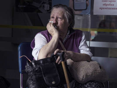 An elderly woman sits in a waiting area, her face conveying worry.