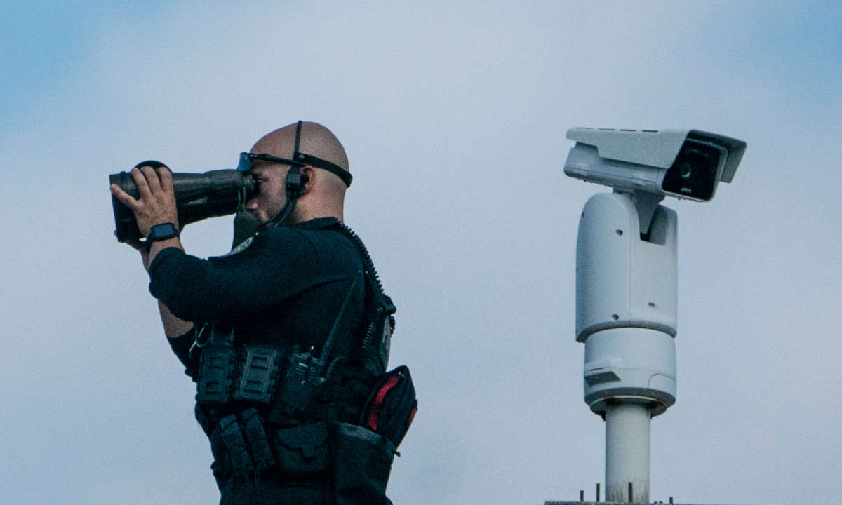 A Secret Service officer looks through huge binoculars while positioned on a roof