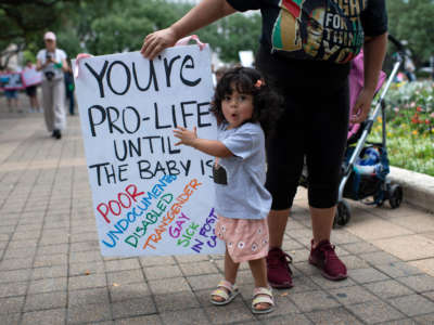 A toddler points to a sign held by her parent that reads 'you're pro-life until the baby is poor / undocumented / disabled / transgender / gay / sick / in foster care" at an abortion rally in Texas