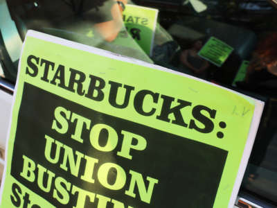 A sign reading: Starbucks: Stop Union Busting, leaned against a car with reflection of protesters in window