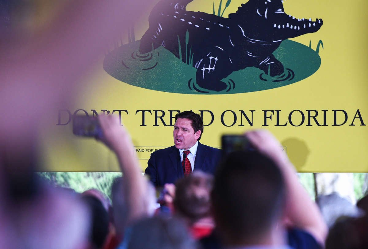 Florida Gov. Ron DeSantis speaks to supporters in front of large yellow 'don't tread on Florida' flag with crocodile illustration