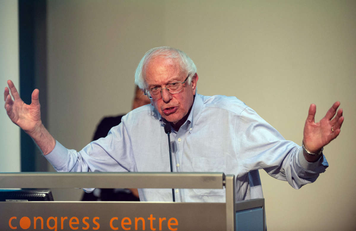 Sen. Bernie Sanders speaks from podium with arms outstretched, in London, England.