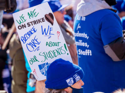 A person in blue cold-weather clothing participates in an outdoor protest while carrying a sign that reads "WE ARE ON STRIKE BECAUSE WE CARE ABOUT OUR STUDENTS"