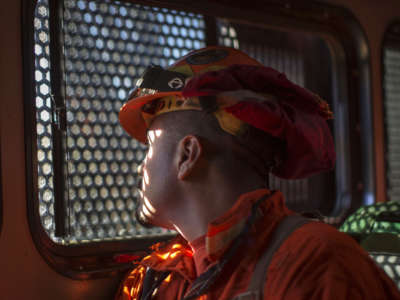 A man in firefighting gear looks out of a bus window covered in perforated metal to prevent escape