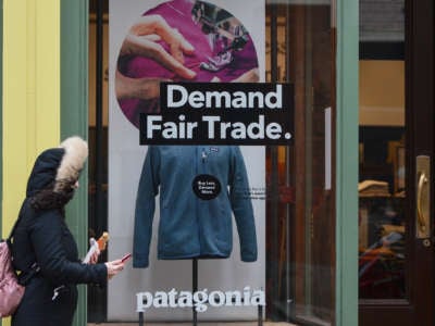 A sign on a Patagonia storefront window reads "Demand Fair Trade." as a person in a coat walks past it
