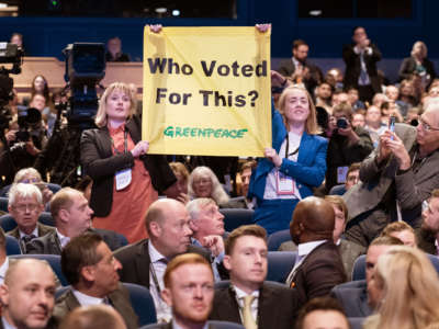 Two people hold up a banner reading "WHO VOTED FOR THIS" beneath which the Greenpeace wordmark is displayed