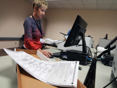 A woman works on a computer as ballots are seen in a box near her