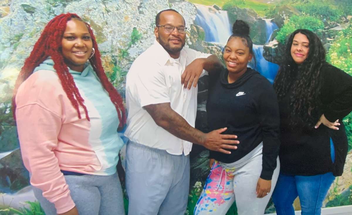 A man stands beside a pregnant woman, whose belly over which he places his hand, as two other women pose next to them in a studio photograph.