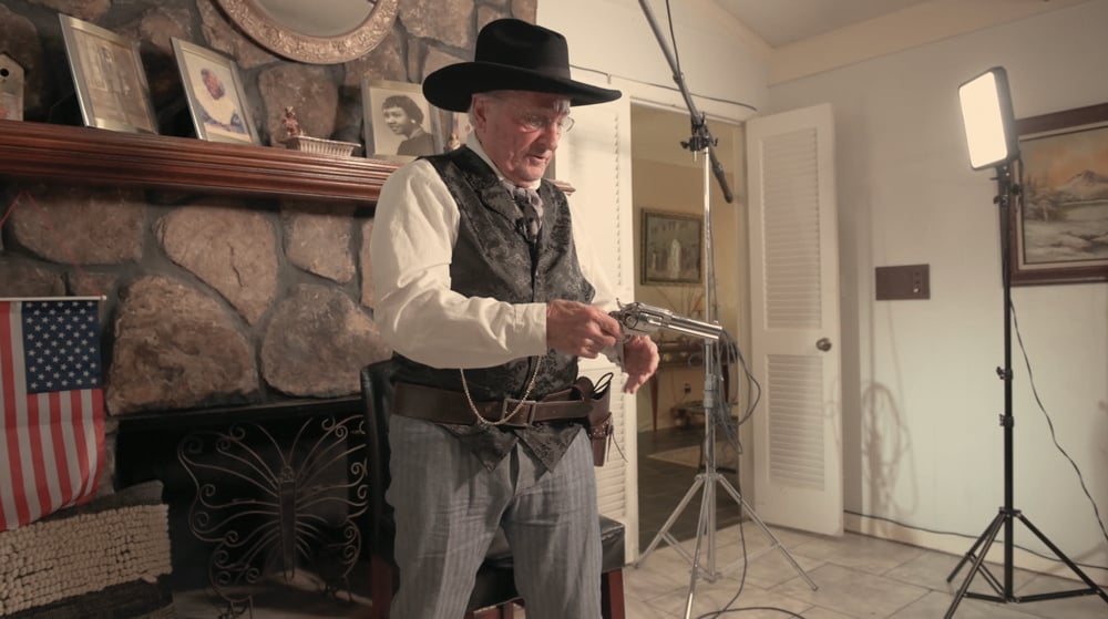 An elderly man in a cowboy hat stands in his living room