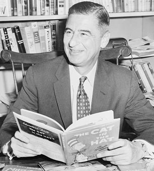 Ted Geisel (Dr. Seuss) seated at a desk covered with his books.