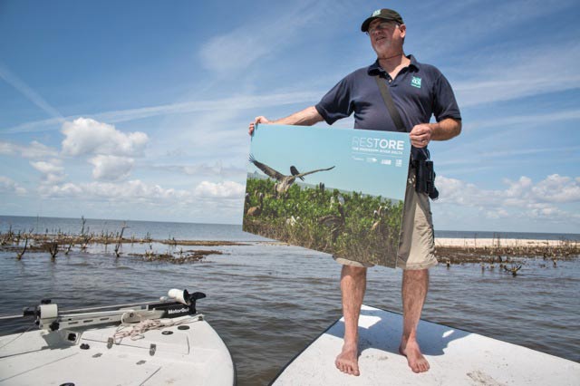 David Muth, Gulf Coast Restoration Director for the National Wildlife Federation holds up a photo of what Cat Island looked like before the BP oil spill, while standing in front of the island on March 31, 2015. (Photo: ©2015 Julie Dermansky)