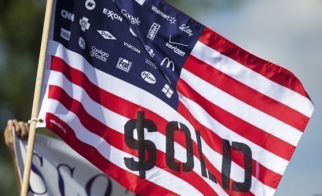 A demonstrator displays a flag decrying corporate spending in American political elections in front of the Waukesha Convention Center on July, 13, 2015, in Waukesha, Wisconsin. (Photo via Shutterstock)