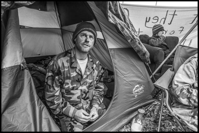 James Cartmill, a veteran and resident of a homeless encampment in Berkeley, California, sits in a tent on the grass median in the middle of Adeline Street. (Photo: David Bacon)