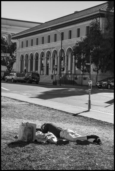 A homeless man sleeps across the street from the Post Office, where the homeless camp used to be. It is now an empty space surrounded by iron bars - the people who used to live there sleep on the grass or elsewhere. (Photo: David Bacon)